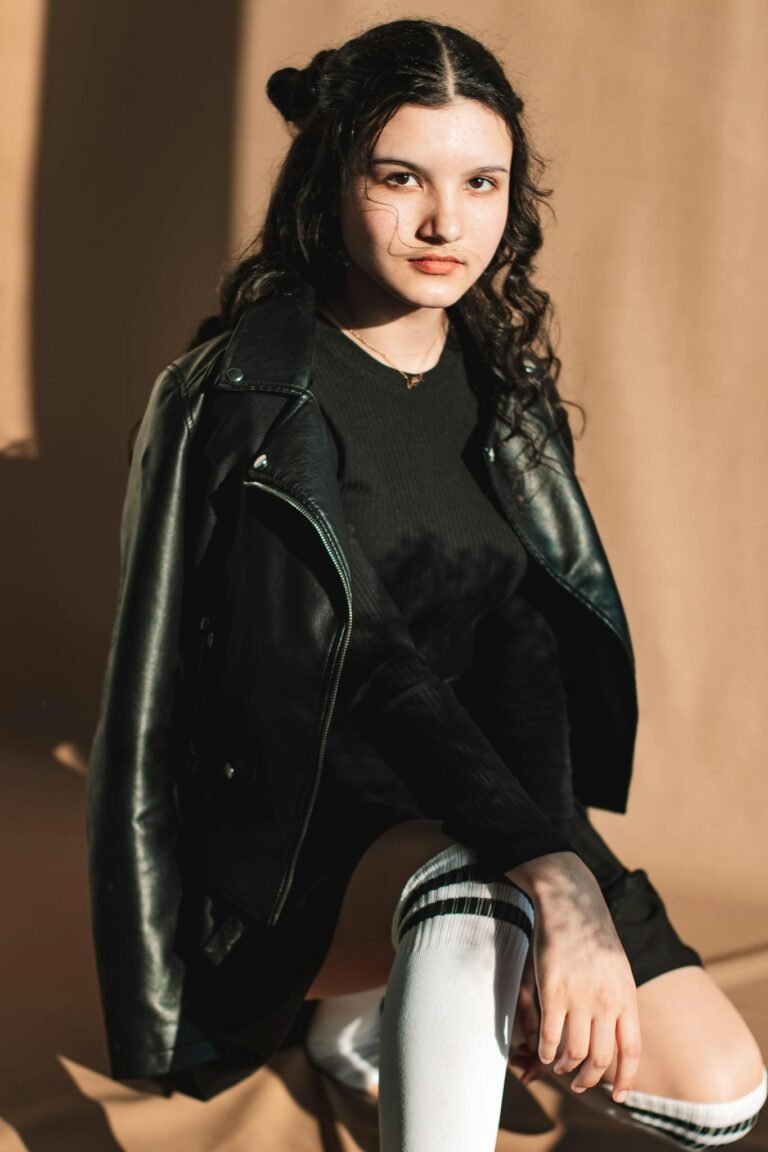 Portrait of a young woman wearing a stylish leather jacket and knee-high socks, posing confidently indoors.